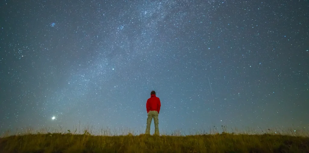 A person stands alone under a massive, starry sky, looking out at the universe and thinking about the real meaning of life