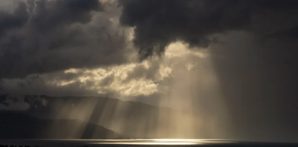 A powerful beam of golden light breaking through dark storm clouds over an open landscape, symbolizing truth emerging through confusion and darkness