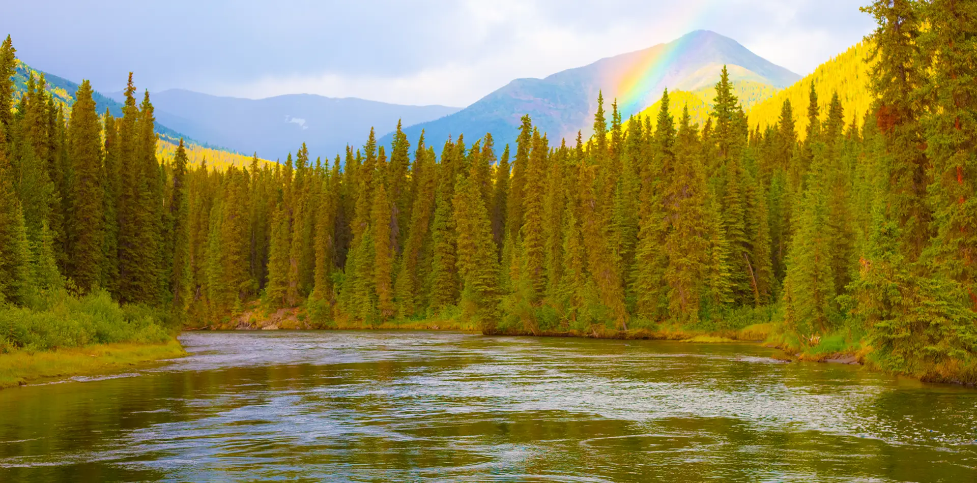 Bright rainbow arching over a flowing river surrounded by colorful natural scenery