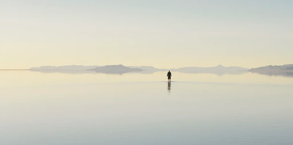 A person standing quietly at sunrise beside a calm shoreline, representing the foundation of love and peace at the beginning of an inner journey.
