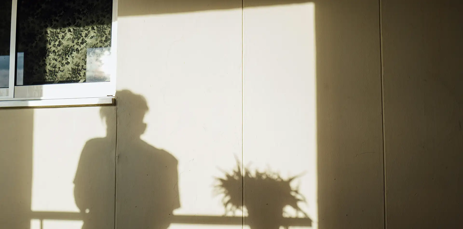 Older person standing quietly at a window in soft morning light, in a calm reflective moment