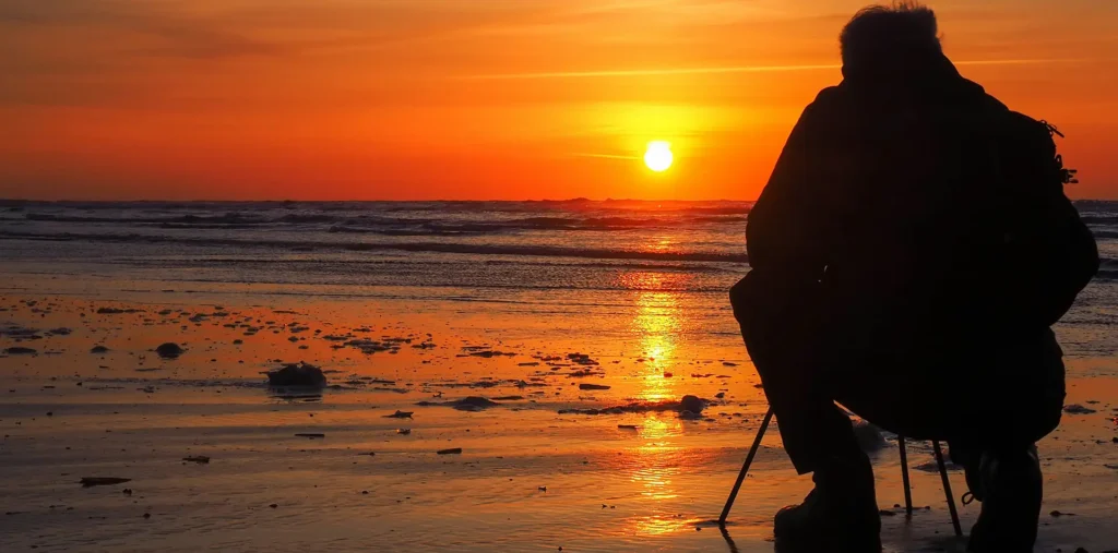 Older person sitting alone watching a golden sunset over the ocean, reflecting on the passing of time