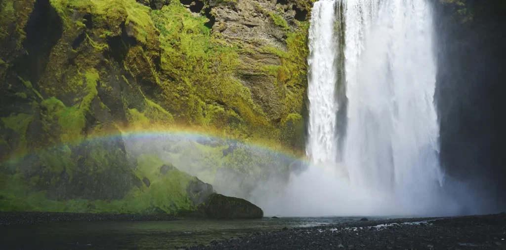 A bright rainbow arching through the mist of a waterfall in a green landscape after a passing storm